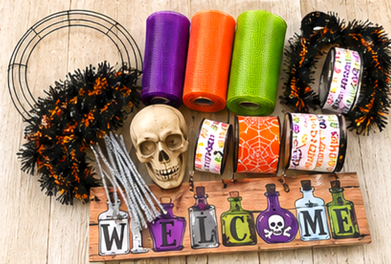 DIY Halloween wreath supplies including a black wire wreath frame, silver pipe cleaners, purple, orange and lime-green deco mesh, Halloween ribbon, spooky skull decorations, and a WELCOME sign.